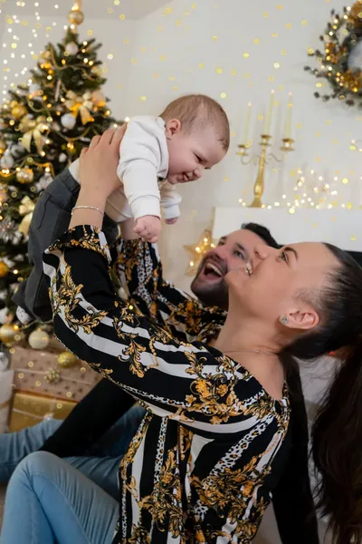Lachende Mama hebt ihr Baby vor dem Weihnachtsbaum hoch