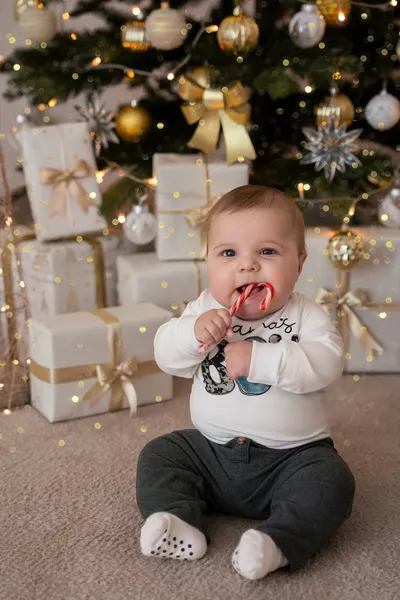 Baby sitzt vor den Geschenken und hält eine rot-weiße Zuckerstange am Mund