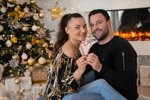 Paar hält gemeinsam eine Zuckerstange vor dem Weihnachtsbaum