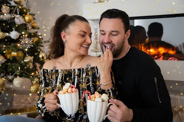 Paar hält Tassen mit Marshmallows, die Frau reicht dem Mann eine Süßigkeit