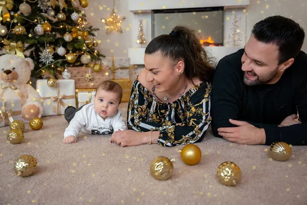 Baby liegt mit beiden Eltern auf dem Bauch zwischen goldenen Weihnachtskugeln