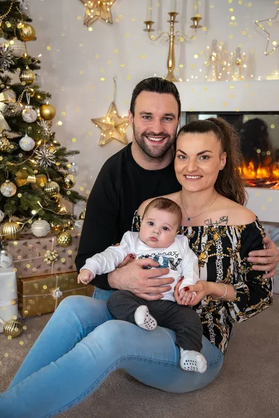 Mama hält das Baby auf dem Schoß, Papa sitzt hinter ihnen am Weihnachtsbaum
