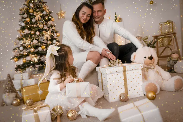 Mädchen wendet sich mit einem Geschenk ihren Eltern am Weihnachtsbaum zu