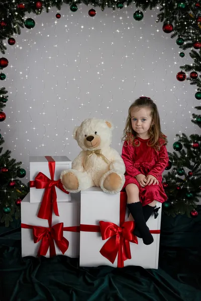 Mädchen im roten Kleid sitzt rechts neben einem Teddy auf weißen Geschenkboxen