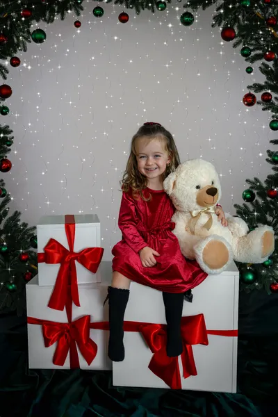 Mädchen im roten Kleid lächelt neben einem Teddy unter dem Tannenbogen