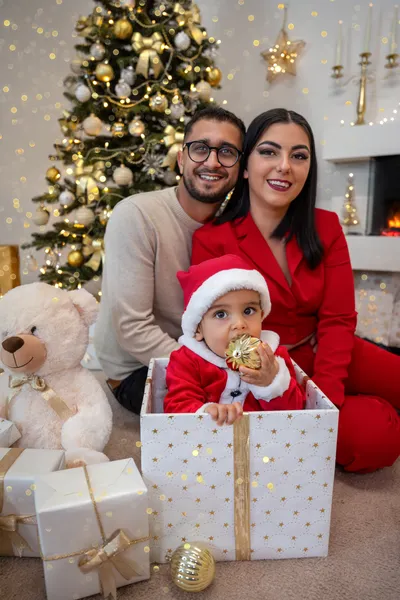 Junge mit Weihnachtsmütze und goldener Kugel sitzt in einer Geschenkbox vor seinen Eltern