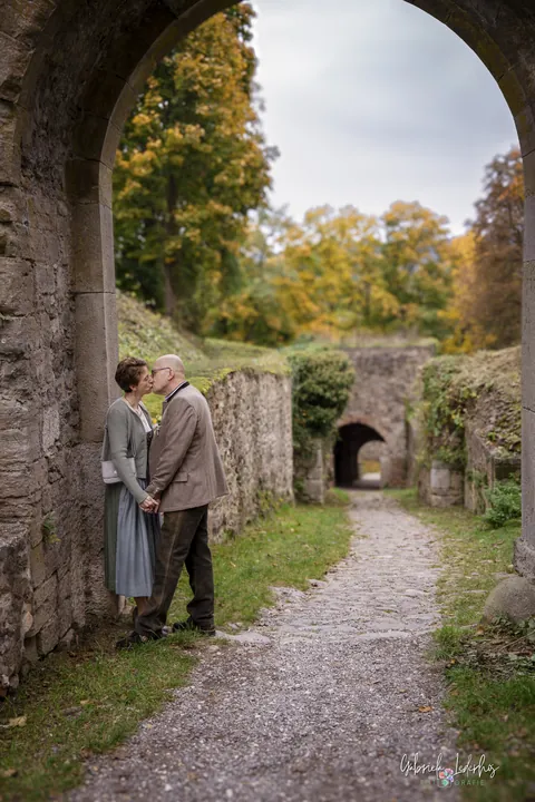 Brautpaar küsst sich im Torbogen einer Burgruine, Herbstlaub im Hintergrund